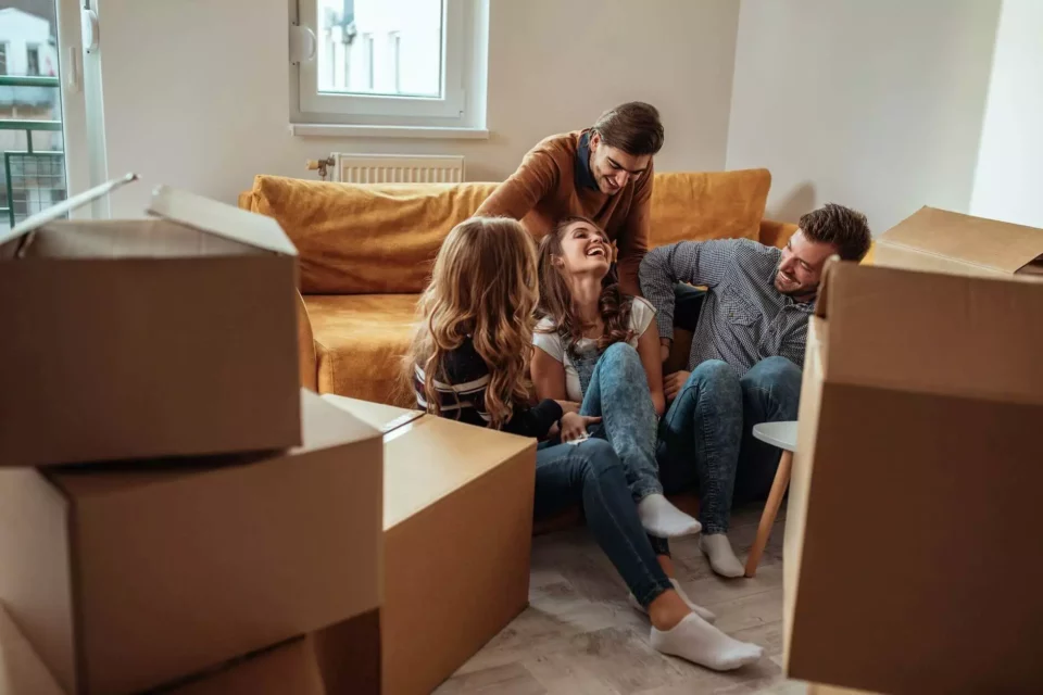Four people laughing in the living room after moving abroad, surrounded by boxes and a yellow sofa 