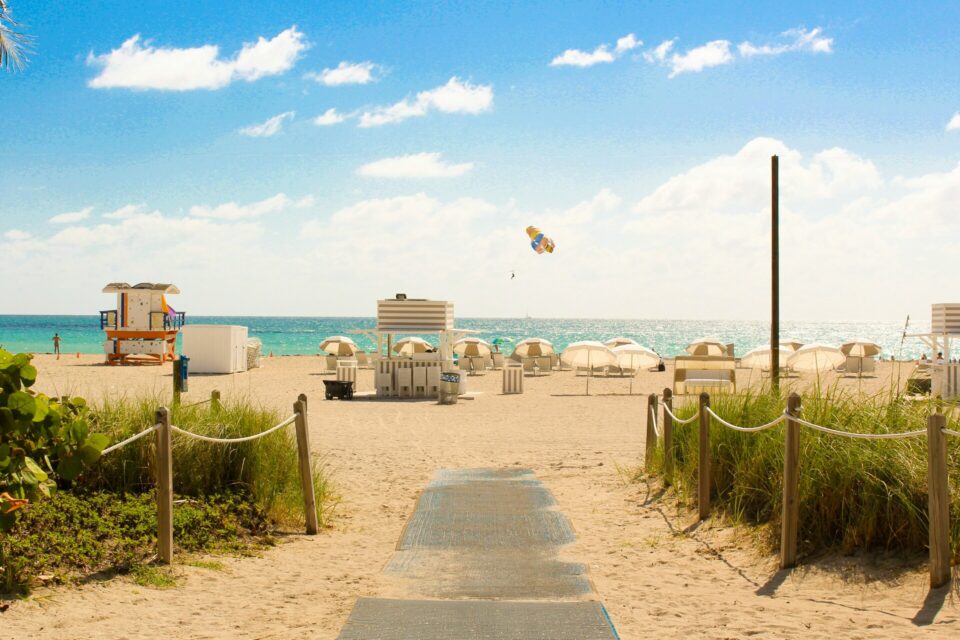 Gray wooden pathway leading to a colorful lifeguard house and beach umbrellas in Miami Beach.